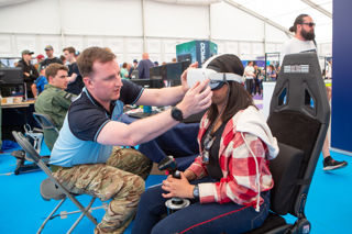 Young girl wearing a virtual reality headset with goggles being adjusted by a man in the Techno Zone at RIAT24.